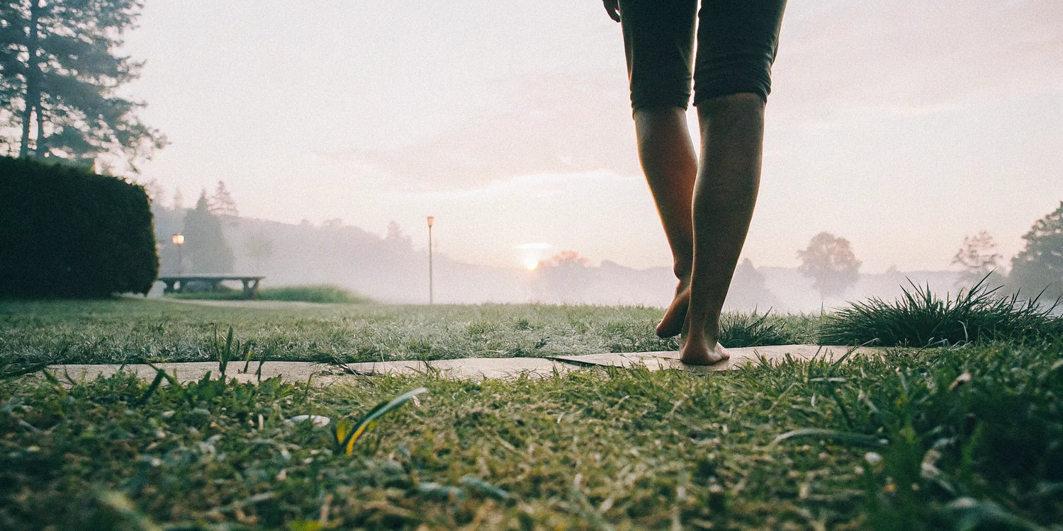 Person walking barefoot on grass at sunrise, grounding with nature