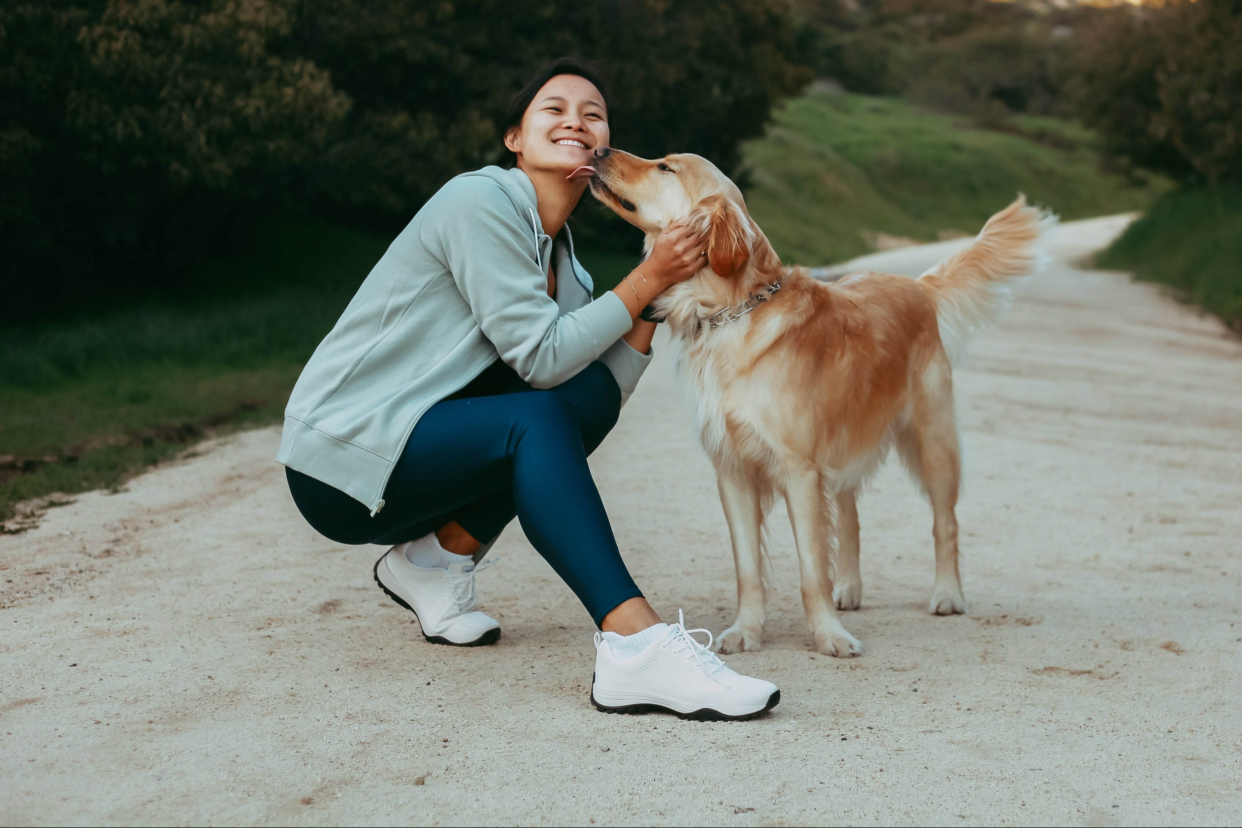 A woman crouches on a dirt path outdoors, smiling as she pets a dog while wearing Harmony 783 Grounding White Sneakers.