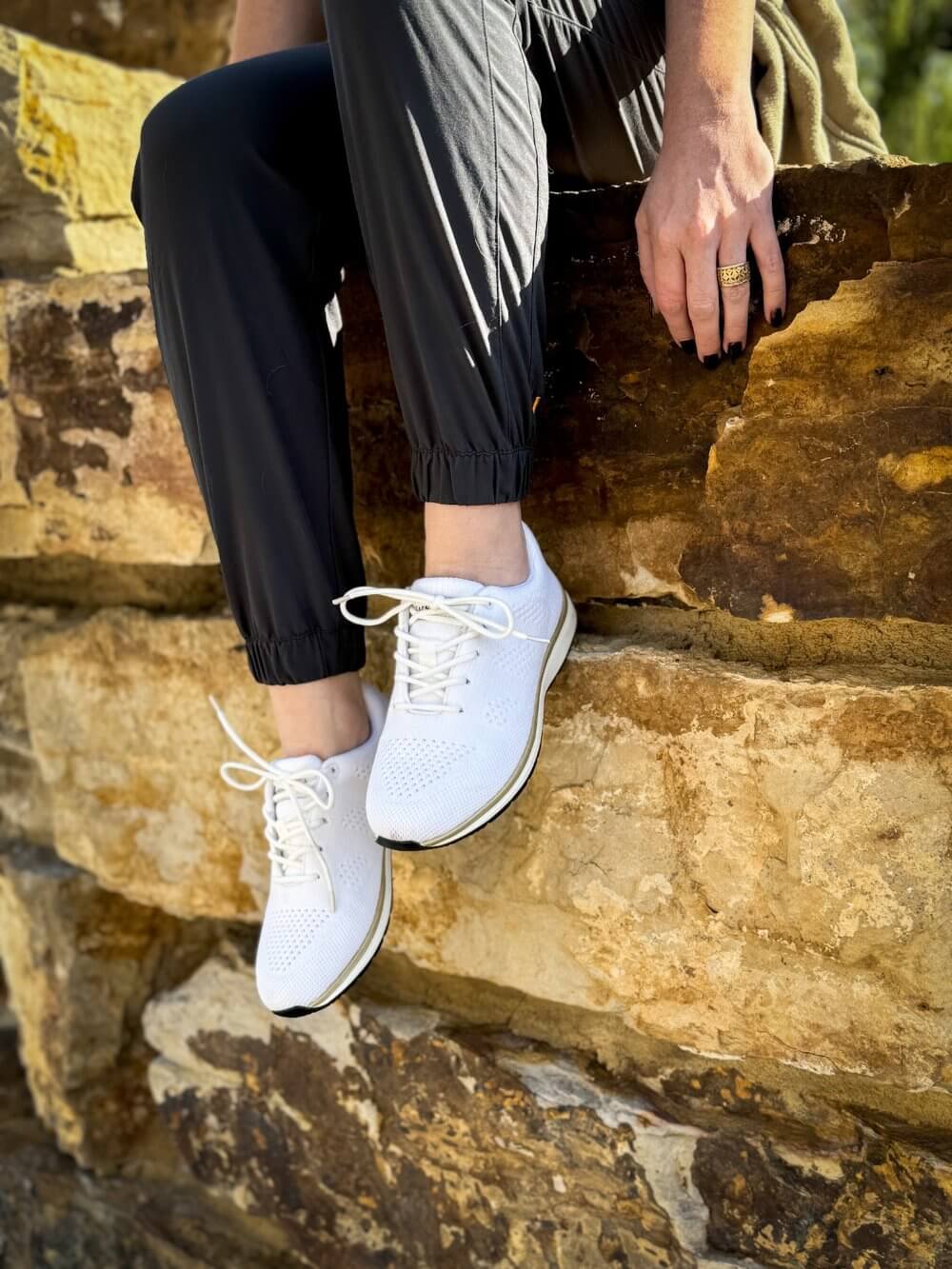 Woman sitting on stone wall with legs hanging over wearing recycled plastic white and gold grounding lace-ups