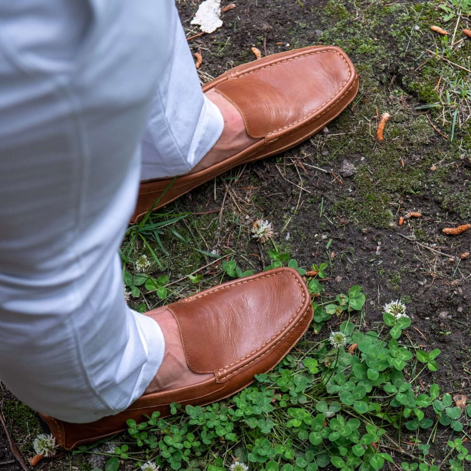 Man wearing brown leather driver and white pants standing on dirt