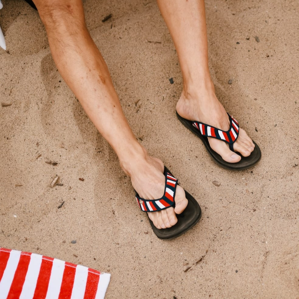 Close up of man sitting on sand at the beach wearing Harmony 783 Red white and blue striped sandal conductive
footbed earthing flip flops-lifestyle