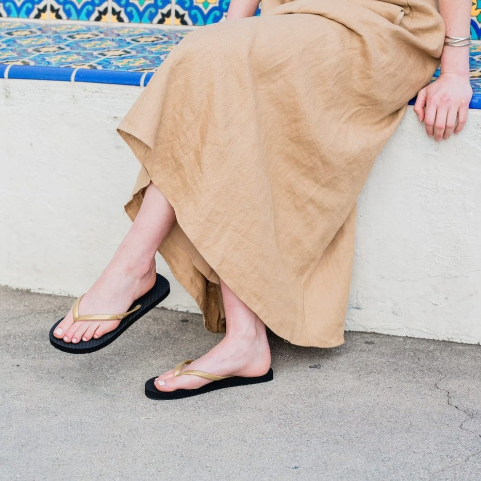 Close up of woman sitting on decorative stairs wearing Harmony 783 black and gold fully conductive footbed earthing flip flops