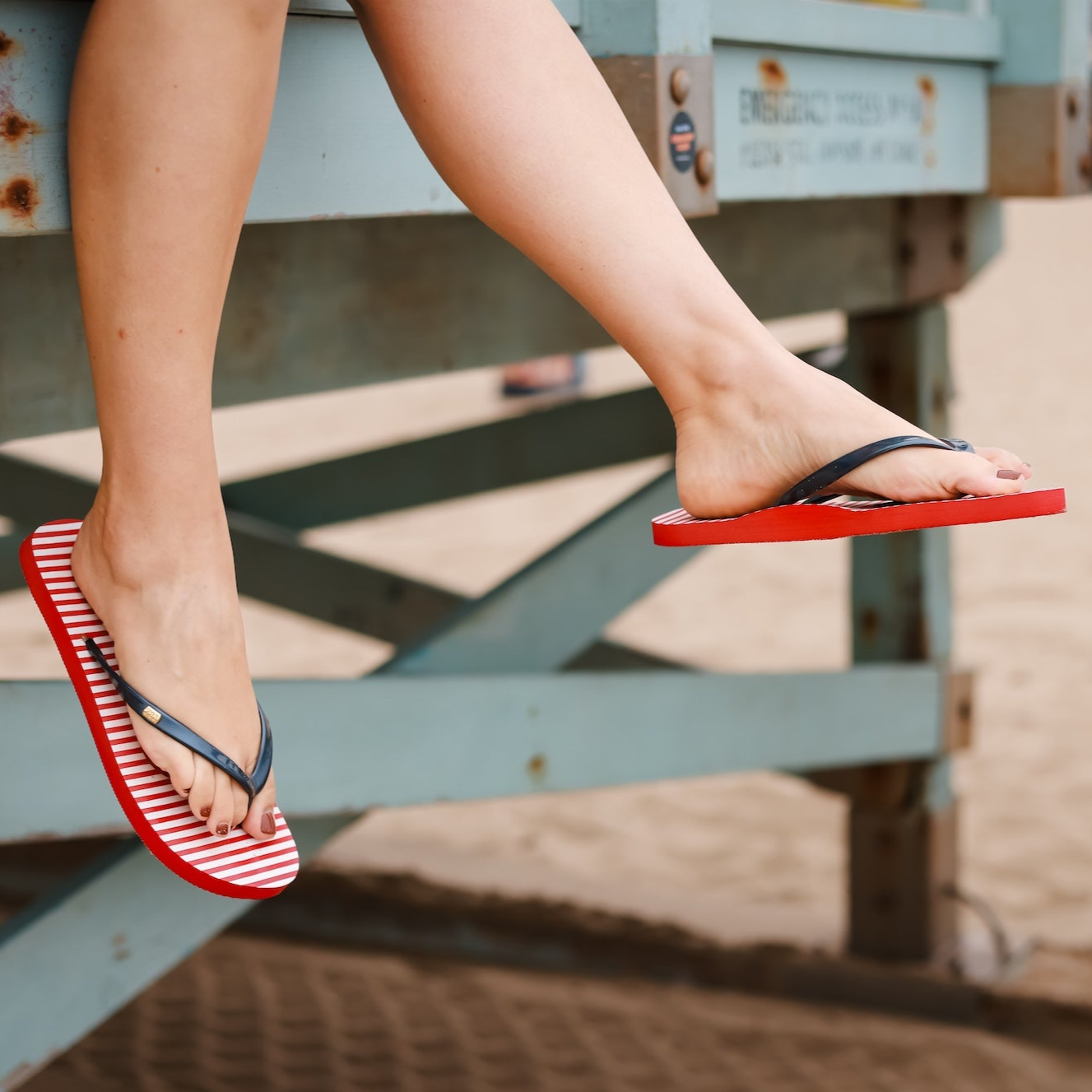 Woman sitting on a pier at the beach with legs hanging over the side wearing red white and blue grounding flip flops