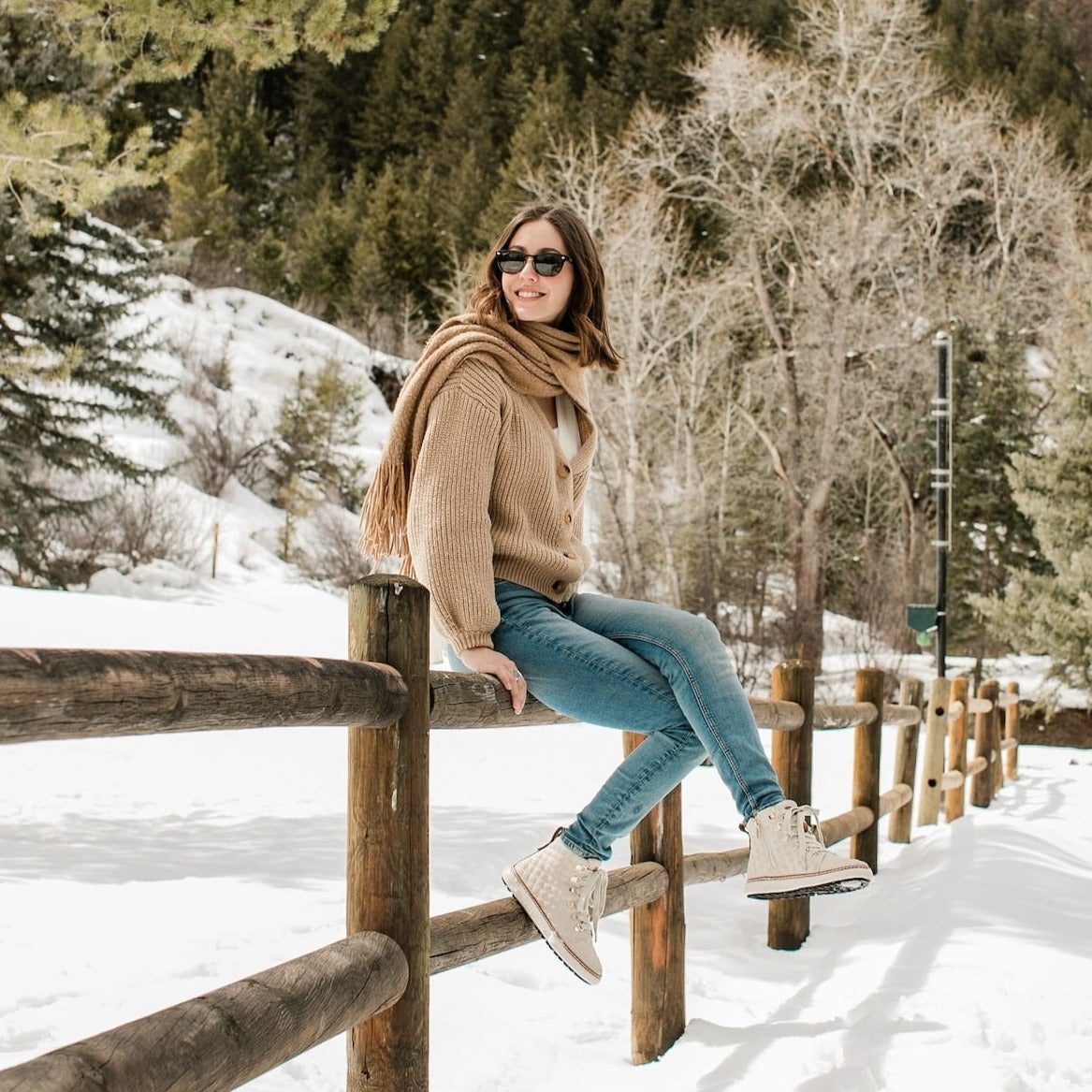 Woman sitting on wood fence in snow wearing gold quilted zip up grounding high-tops 