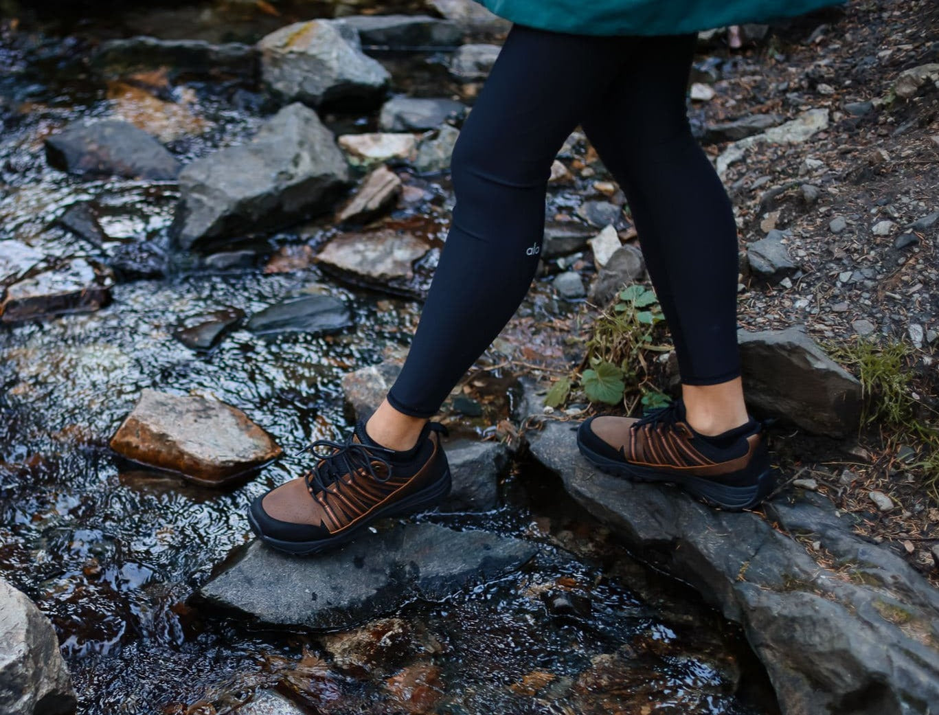 Woman walking on stones near a river wearing black leggings and brown leather grounding trail shoes